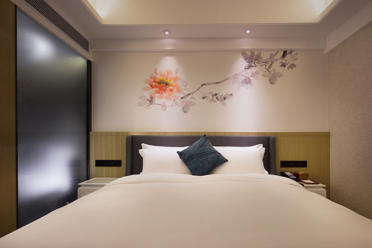 Ein Hotelzimmer mit einem großen Bett, einem Nachttisch, einem an der Wand befestigten Telefon und einem floralen Wandbild.