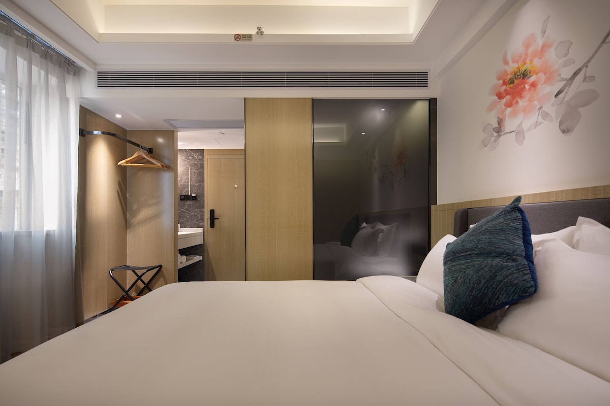 Moderni hotellihuone, jossa on suuri sänky, erillinen vaatehuone, suihkulla varustettu kylpyhuone ja suuri ikkuna, jossa on verhot.