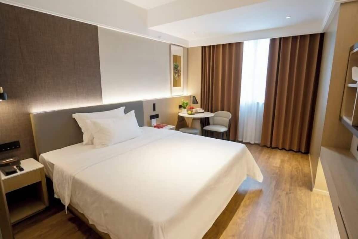 A hotel room with a large bed, a bedside table, a small desk with a chair, and a window with curtains.