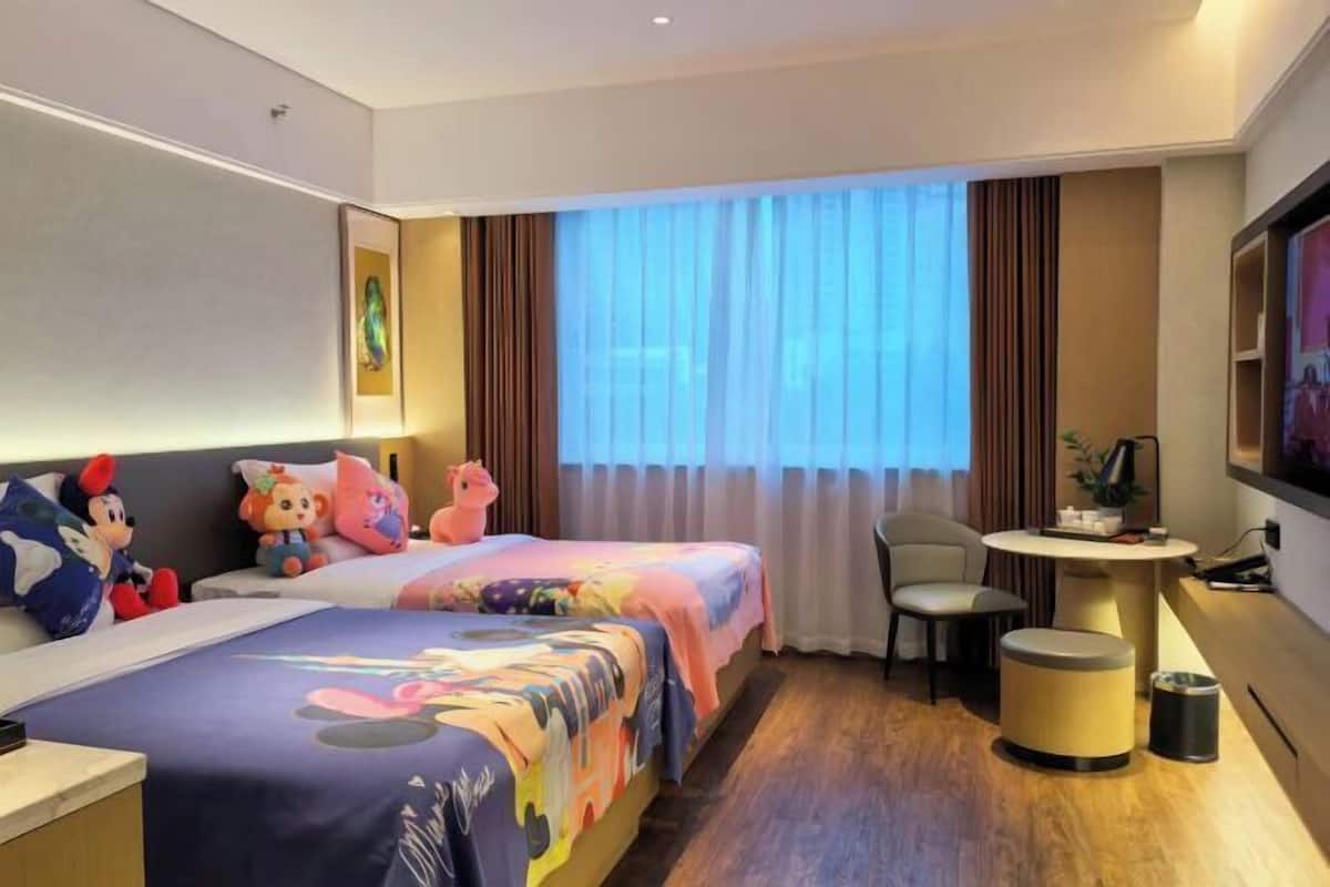 A hotel room with two beds, plush toys, a small table, a chair, and a TV.