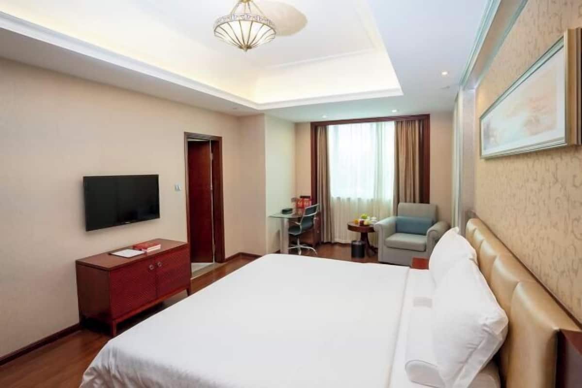 A hotel room with a large bed, a TV, a desk with a chair, a small table with a chair, and a window with curtains.
