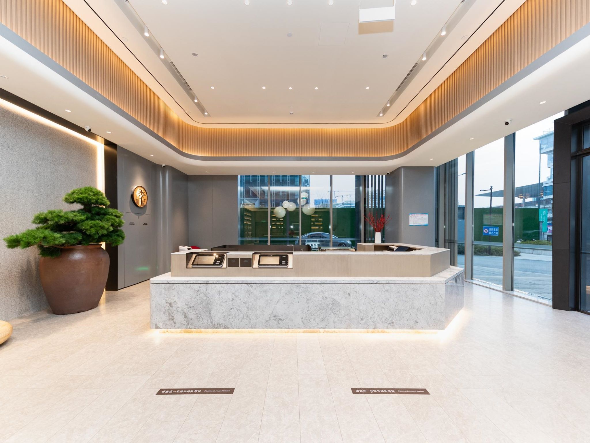 A reception desk with a modern design, a large potted plant, and a view of the outside through large windows.