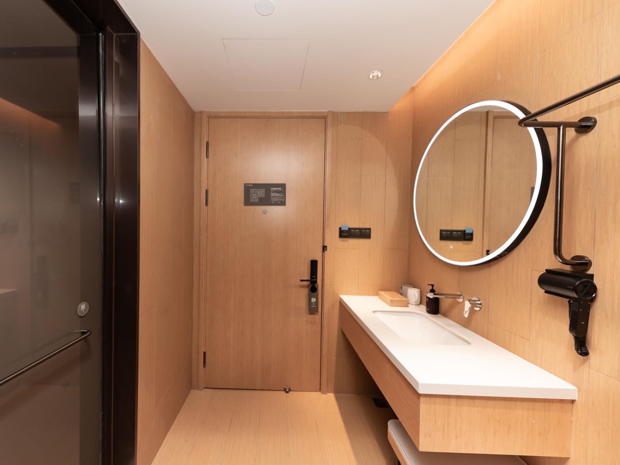 A modern bathroom with a wooden door, a white sink, a round mirror, and a handheld hair dryer.