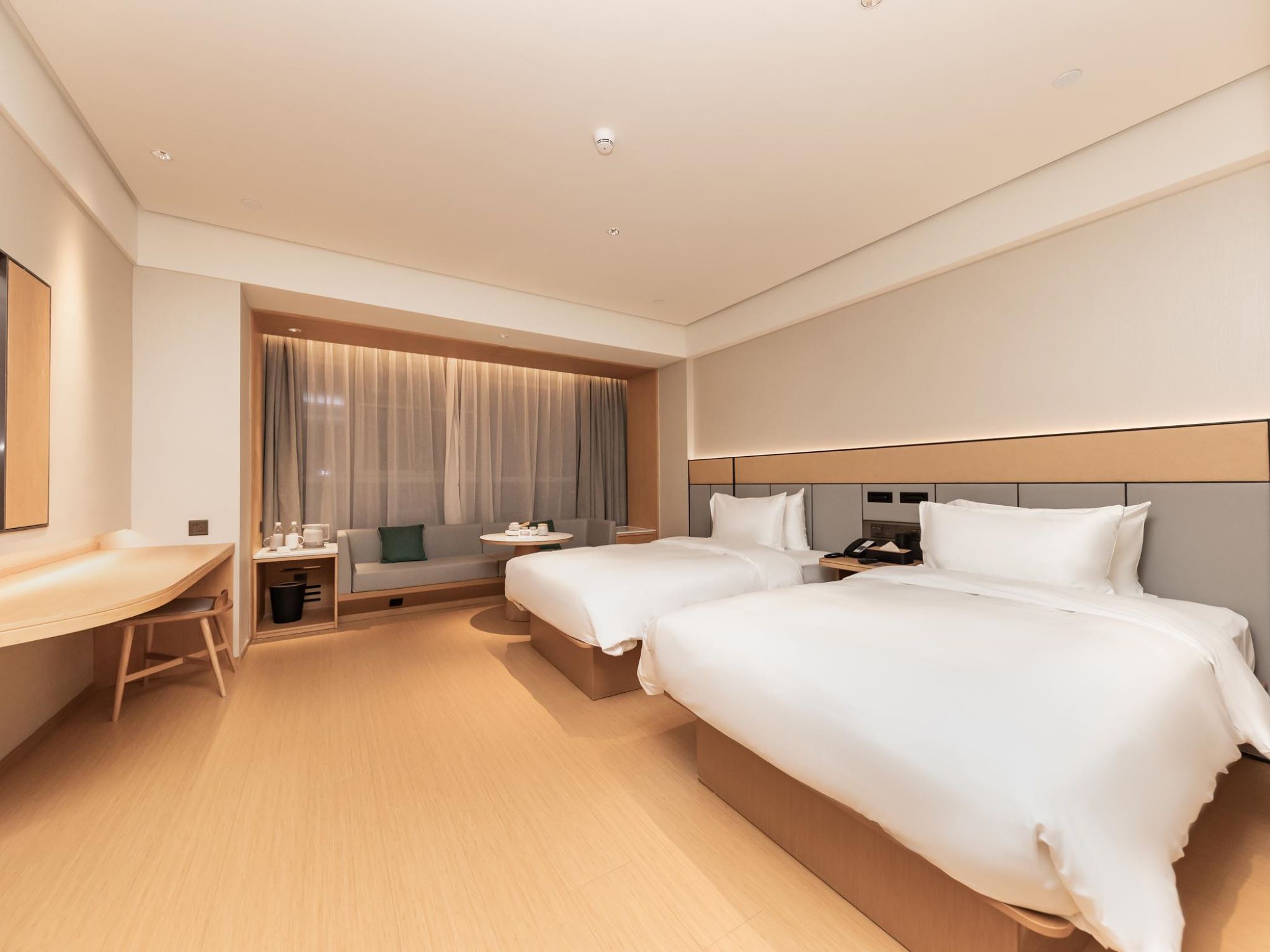 A hotel room with two beds, a desk, a sofa, and a large window with curtains.