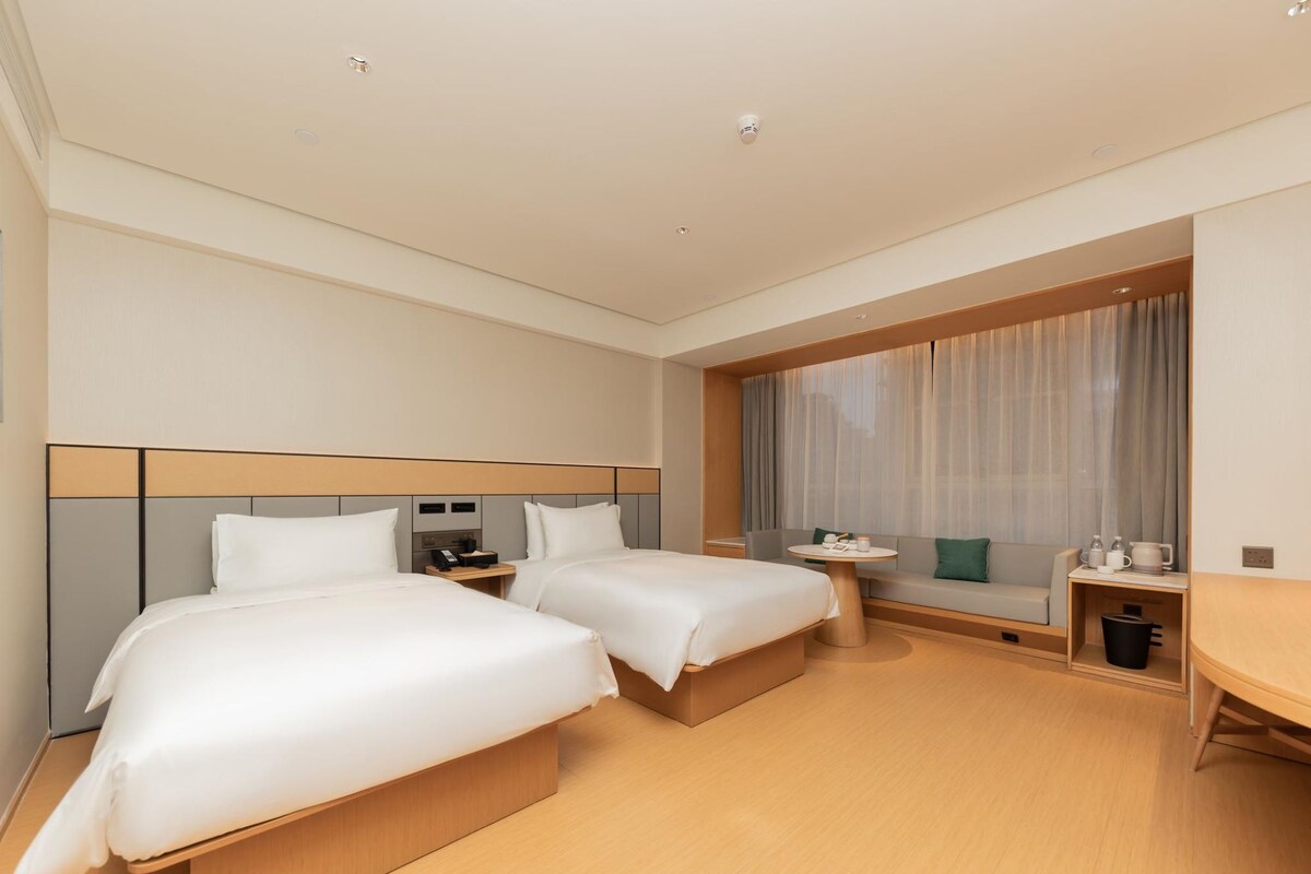 A hotel room with two beds, a small table, a sofa, and a window with curtains.