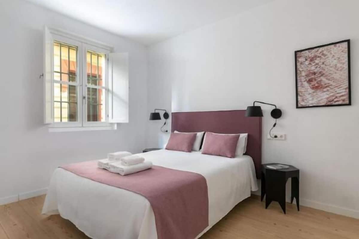 A bedroom with a bed, bedside tables, a window, and a wall-mounted lamp.