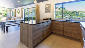 A modern kitchen with a large island, wooden cabinets, and a view of a residential area. - Spa Pool, Sea and Stunning Views | Cass Bay Escape (Cass Bay)