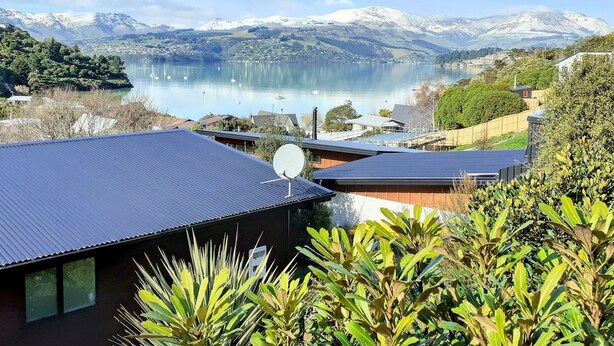 A modern building with a metal roof, surrounded by lush greenery and a view of a lake with boats and snow-capped mountains in the background. - Spa Pool, Sea and Stunning Views | Cass Bay Escape (Cass Bay)