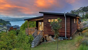 A modern wooden cabin with a balcony, situated on a hillside with a view of the ocean and other houses. - Spa Pool, Sea and Stunning Views | Cass Bay Escape (Cass Bay)