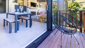 A modern living room with a fireplace, a dining area with a table and chairs, and a balcony with chairs. - Spa Pool, Sea and Stunning Views | Cass Bay Escape (Cass Bay)