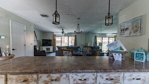 A kitchen with a granite countertop, white cabinets, and pendant lighting. There is a dining area with a sofa and table, and a large window providing natural light.