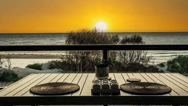 Een houten terras met geweven matten en een plant in pot, met uitzicht op een strand bij zonsondergang.