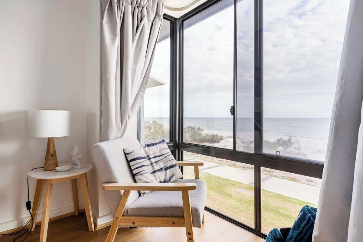 Une chambre moderne avec une grande fenêtre, une chaise en bois, une petite table avec une lampe et une vue sur la mer.