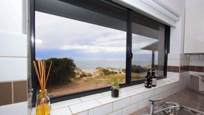 Een keuken met een groot raam dat uitkijkt op een strand, een spoelbak en een vensterbank met een plant en twee flessen.