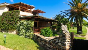 A two-story house with a stone wall, a covered patio, and a garden with palm trees. - Blue&green House With Shared Pool (Golfo Aranci)