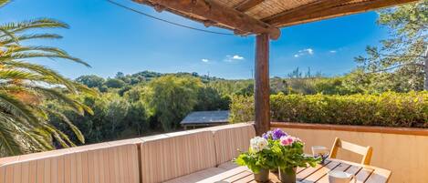Ein Holztisch mit zwei Tassen, einem Blumentopf und einer Bank unter einer Holzpergola mit Blick auf grüne Vegetation.