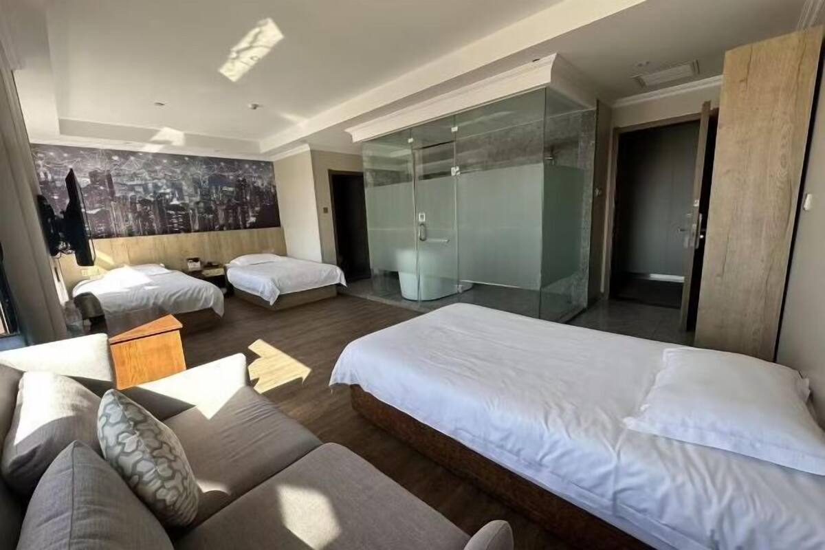 A hotel room with two beds, a sofa, a glass wardrobe, and a cityscape mural on the wall.