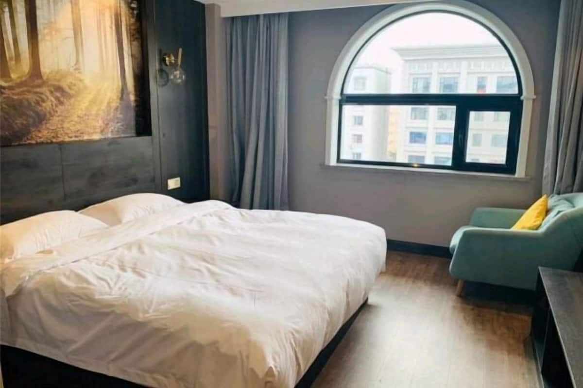 A hotel room with a bed, a sofa, a window with a view of buildings, and a painting on the wall.