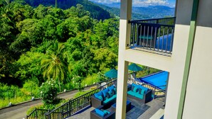 A balcony with a view of a lush green landscape and a pool area with blue furniture.