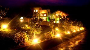 A two-story house with a balcony, surrounded by well-lit gardens and a pathway.