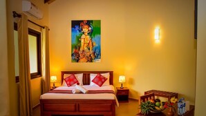 A hotel room with a wooden bed, two bedside lamps, a painting on the wall, and a small table with fruit and water bottles.