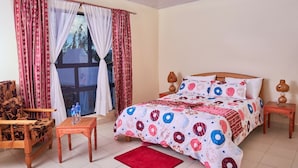 A bedroom with a large bed, a wooden chair, a small table, and a window with curtains. - Galaxy Inn - Athi River (Athi River)