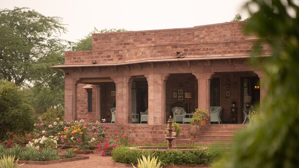 A brick building with a covered patio area, a fountain, and a garden with various plants and flowers.