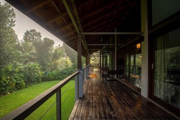 A covered outdoor area with a wooden deck, a glass wall, and a view of a well-maintained garden.