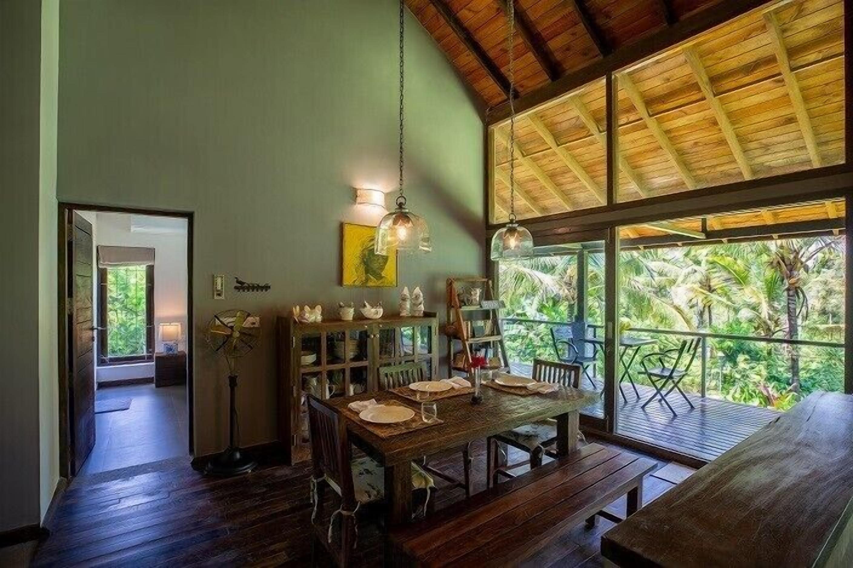 A dining area with a wooden table, chairs, and a fan. There is a balcony with a view of greenery.