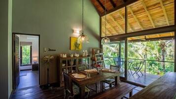 A dining area with a wooden table, chairs, and a fan. There is a balcony with a view of greenery.