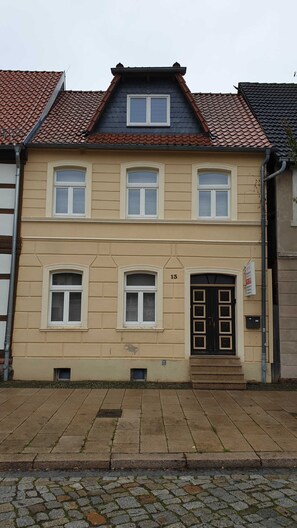 Ein zweistöckiges Gebäude mit einer symmetrischen Fassade, das einen zentralen Eingang mit einer schwarzen Tür und eine kleine Treppe davor besitzt.