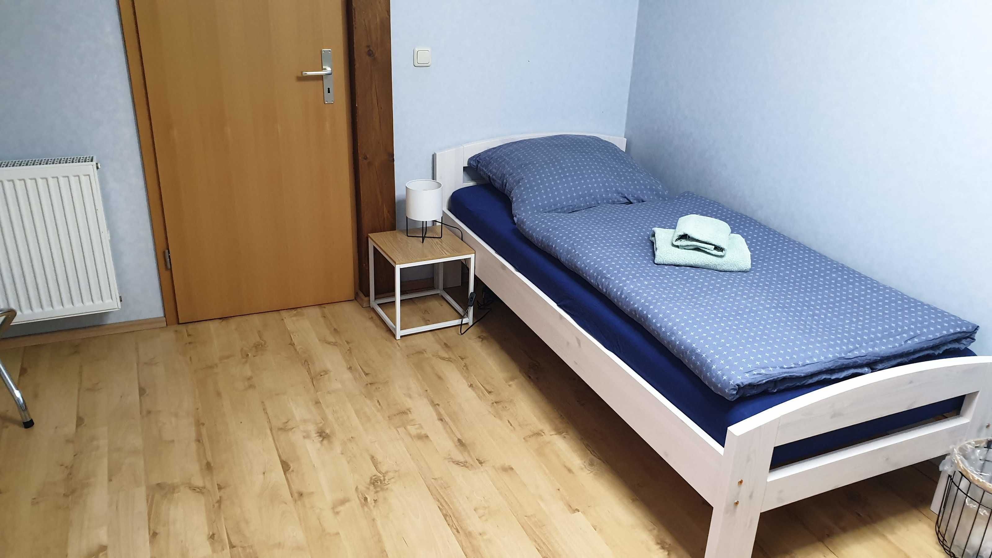 Une petite chambre avec un lit simple, un plancher en bois, une table de chevet avec une lampe et une porte dotée d’une poignée.