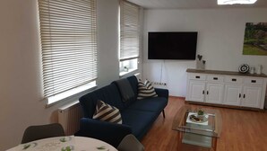 Sala de estar com um sofá azul, uma mesa de centro de vidro, um armário branco e uma televisão de tela plana fixada na parede.