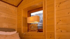 Ein Zimmer einer Holzhütte mit einem Bett, einem Fenster mit Blick auf schneebedeckte Bäume und einer Lampe auf einem Regal.