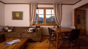 Ein gemütliches Wohnzimmer mit einer Couch, einem Esstisch mit Stühlen, einem Fenster mit Vorhängen und einem Bild an der Wand.
