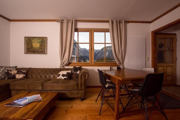 A cozy living room with a sofa, a dining table with chairs, a window with curtains, and a painting on the wall.