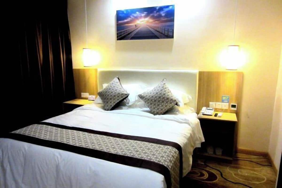 Hotellihuone, jossa on suuri sänky, kaksi yöpöytää valaisimineen, sängynpääty ja taulu seinällä.