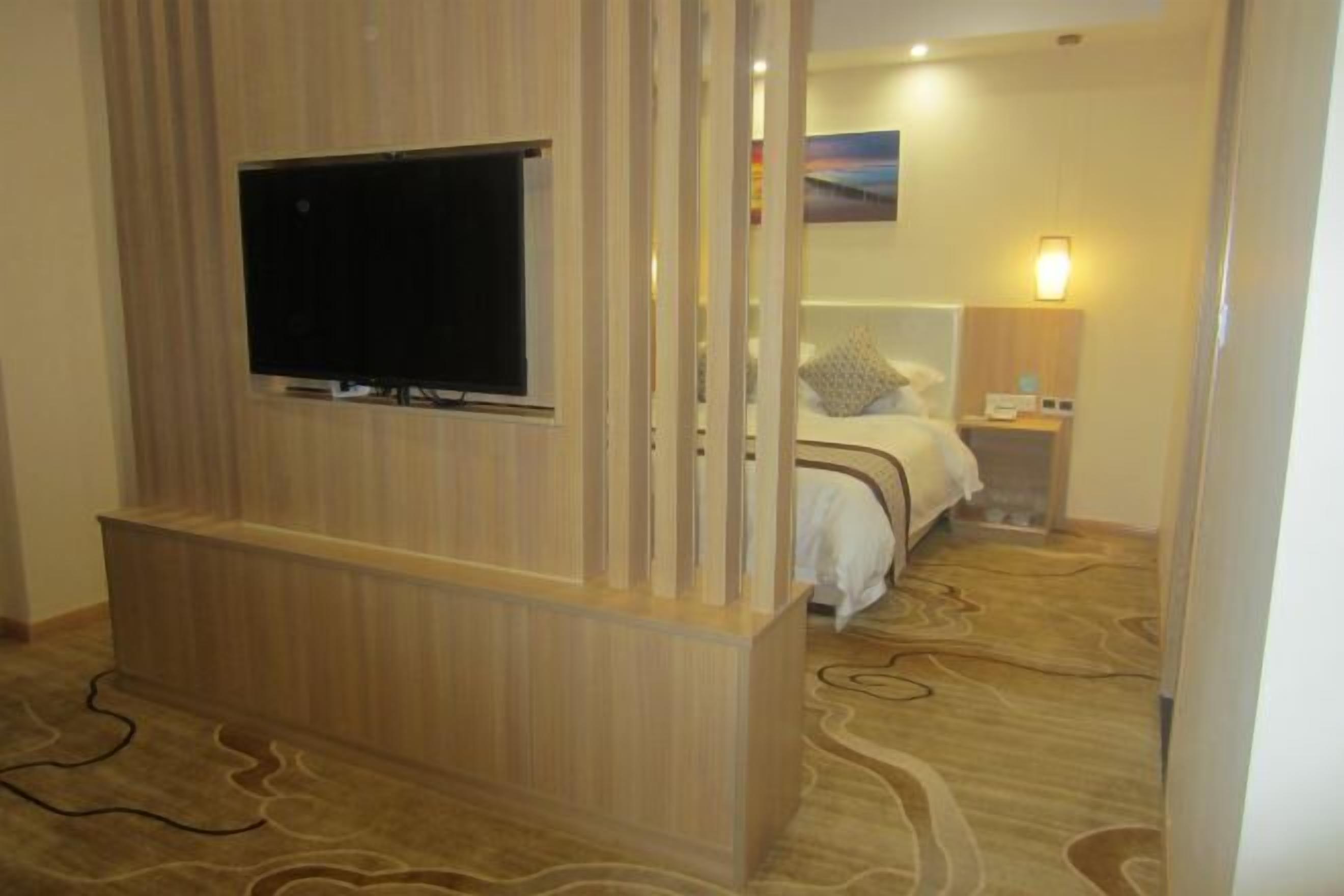 A hotel room with a TV mounted on a wooden panel, a bed with a headboard, a bedside table with a lamp, and a carpet with a wavy pattern.