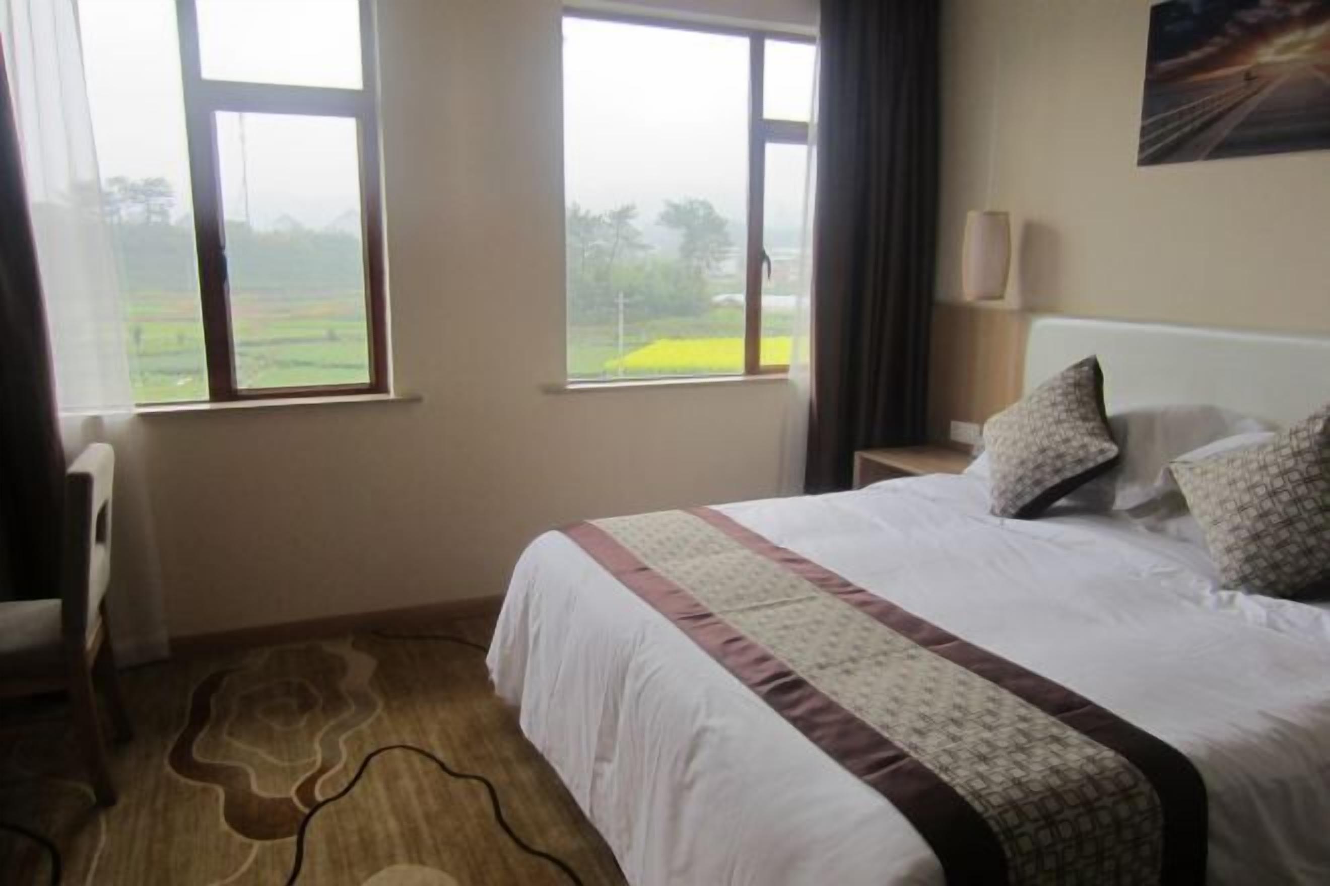 A hotel room with a bed, two windows, a bedside table, and a lamp.