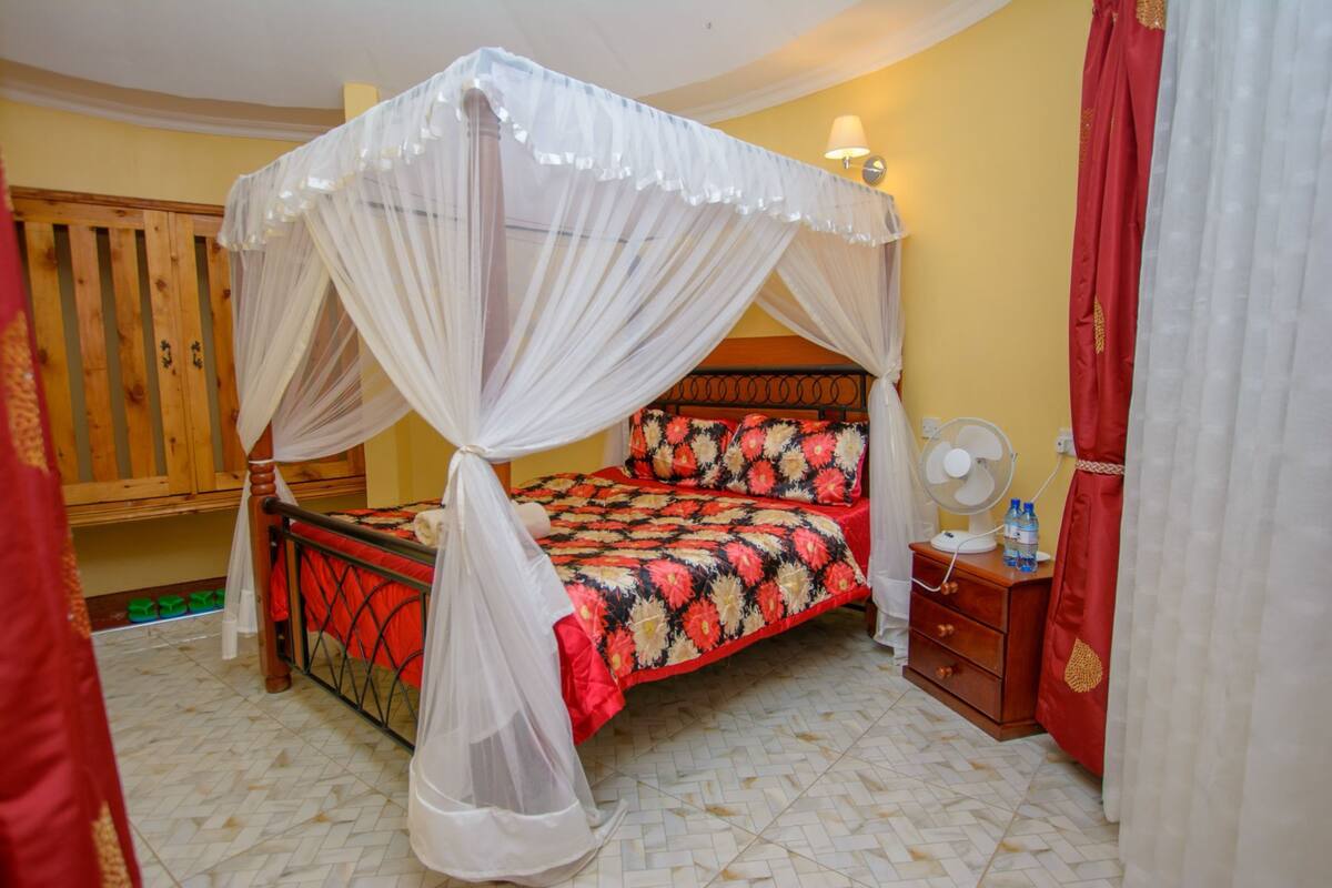 A bedroom with a four-poster bed, a wooden wardrobe, a bedside table with a fan, and a red curtain.