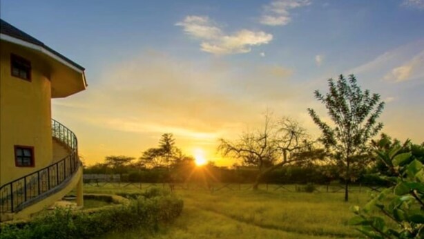 A yellow building with a curved balcony, surrounded by a grassy field and trees, with the sun setting in the background. - Lomanira Splendour (Mogotio)