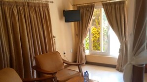 A hotel room with two wooden chairs, a small wooden table with a bottle and remote control, beige walls, and a window with curtains. - Lomanira Splendour (Mogotio)