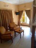 Habitación de hotel con dos sillas de madera, una pequeña mesa de madera con una botella y un control remoto, paredes color beige y una ventana con cortinas.