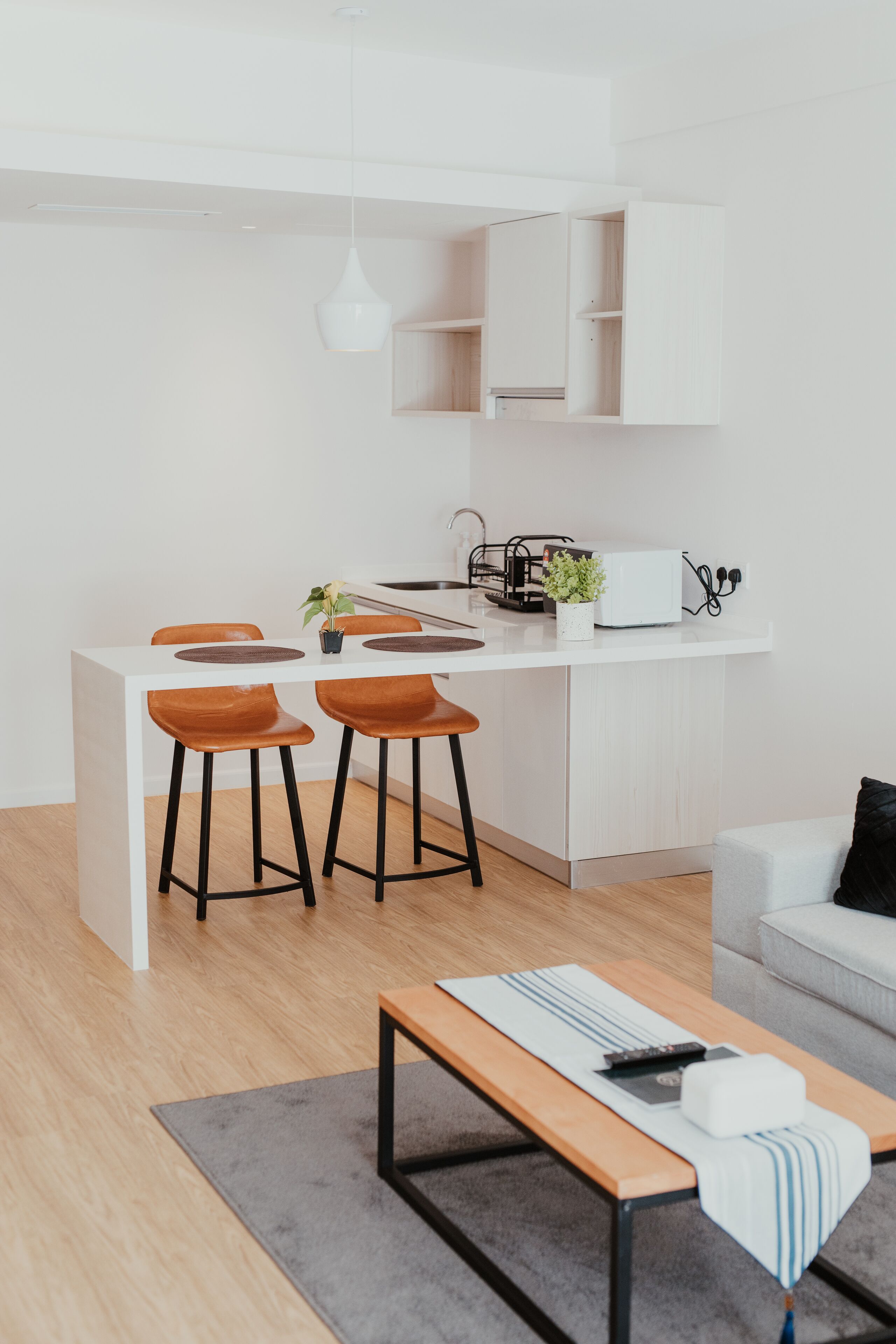 Une cuisine moderne avec des meubles blancs, un coin repas avec des chaises en bois, un petit îlot central et un canapé gris dans un espace de vie lumineux.