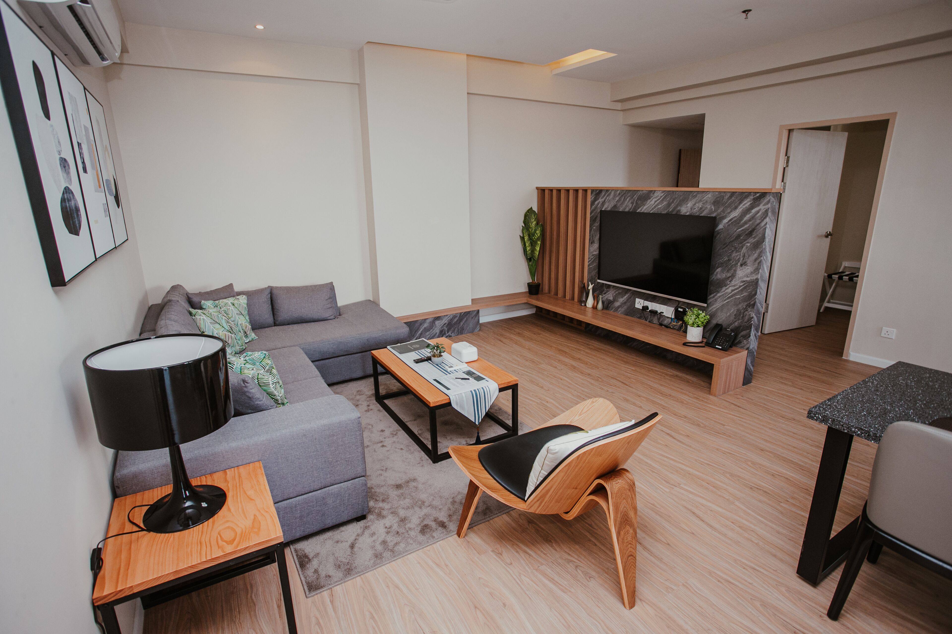 A modern living room with a grey sofa, a wooden coffee table, a black floor lamp, a wooden chair, a TV on a marble wall, and a small table with a plant.