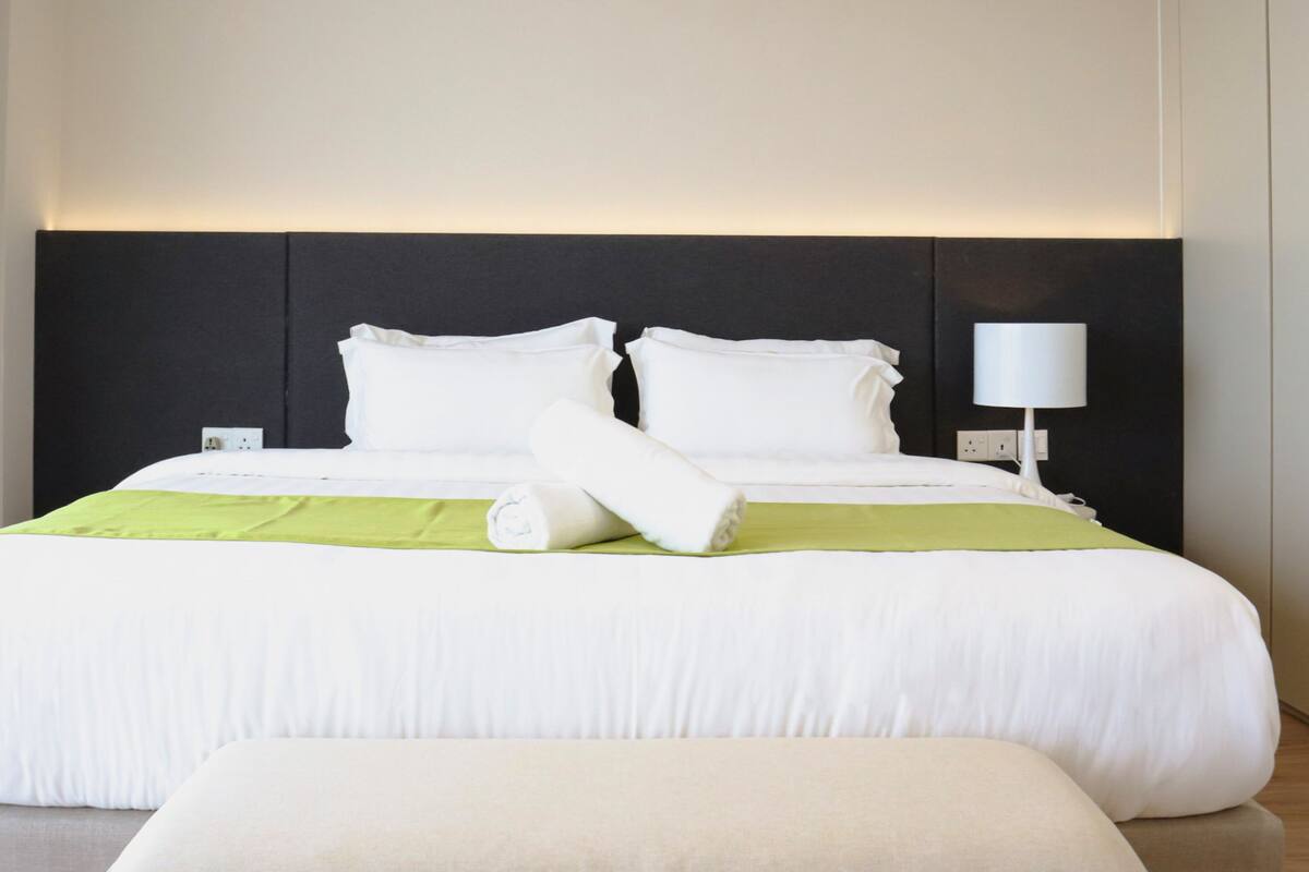 A neatly made bed with white linens and a green accent, a bedside lamp, and a power outlet on the wall.