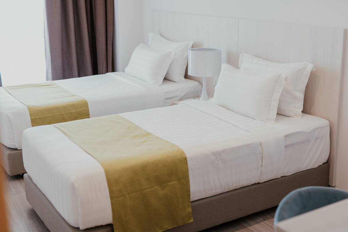 Two twin beds with white bedding and a yellow accent, a bedside table with a lamp, and a view of a window with curtains.