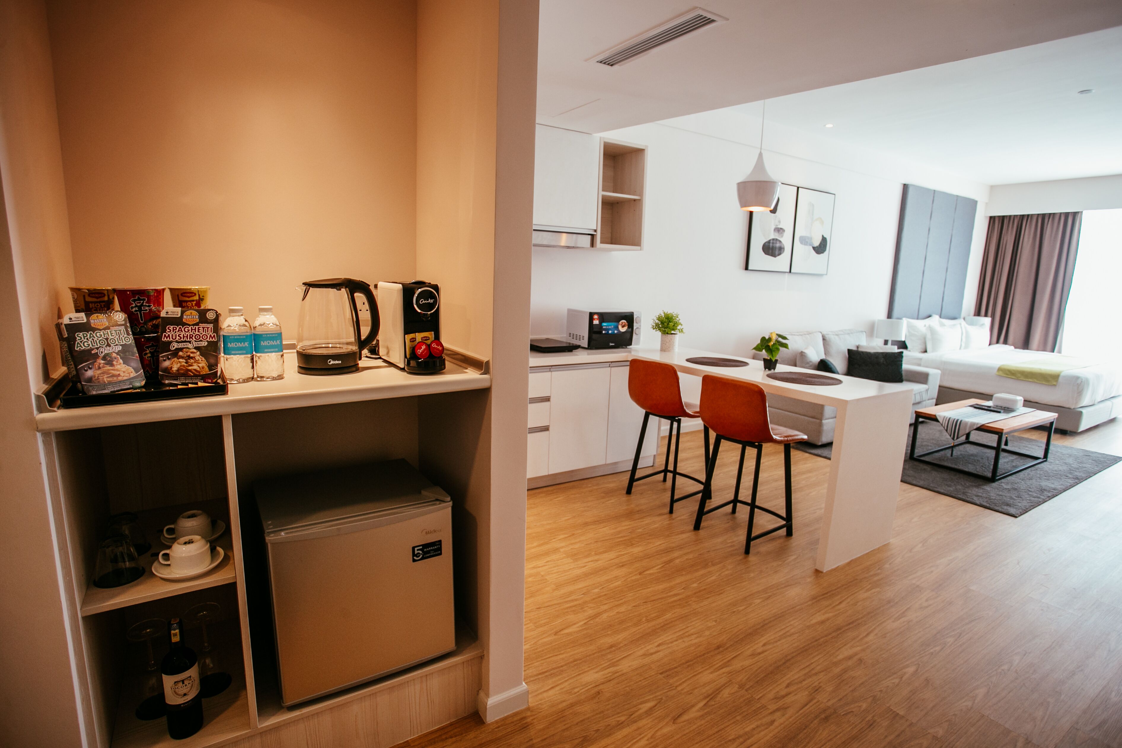 A modern hotel room with a kitchenette, a dining area with two red chairs, a small refrigerator, and a coffee maker.