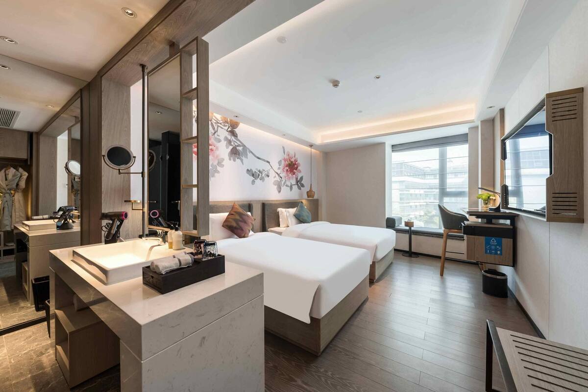 A modern hotel room with a large mirror, a vanity with a sink, two beds, a desk, and a large window offering a city view.