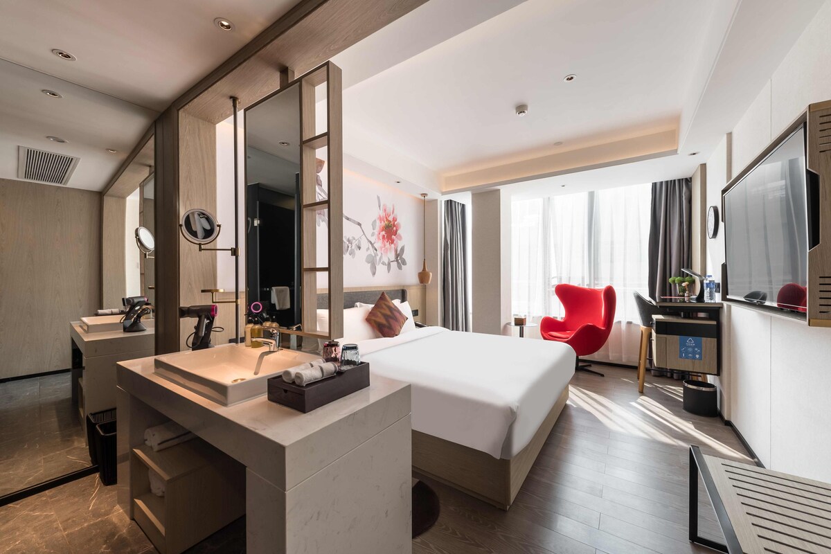 Ein Hotelzimmer mit einem großen Bett, einem Schreibtisch mit Computer, einem roten Sessel und einem Badezimmer mit Waschbecken und Spiegel.